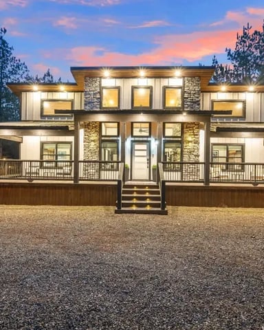 Modern two-story house with stone and metal siding, lit up at dusk, surrounded by tall pine trees