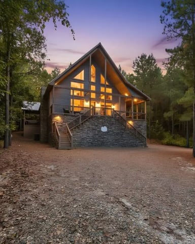 Modern cabin with stone base and large windows lit at dusk, surrounded by tall pine trees and gravel driveway.