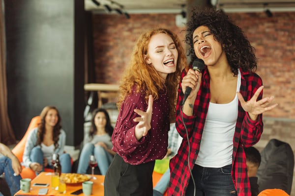 Two women singing karaoke with microphone at indoor party with friends watching, brick wall background