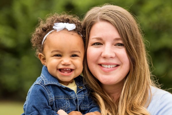 Woman and toddler smiling outdoors with blurred green foliage background