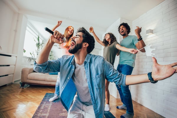Group of friends singing and dancing in a bright living room with white brick walls and wooden floors