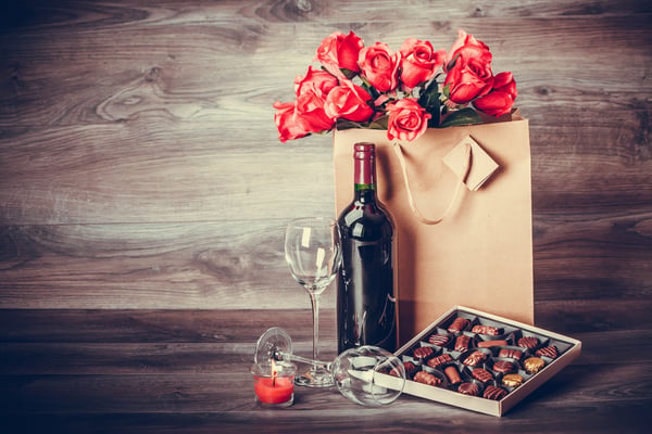 Red roses in kraft paper bag, wine bottle, wine glass, chocolates, and candle on wooden surface