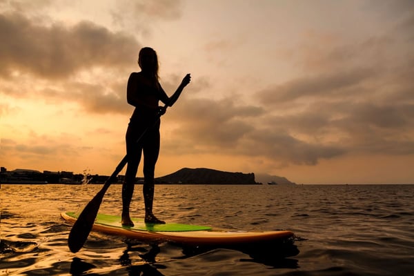 Silhouette of person stand-up paddleboarding at sunset with mountains visible across the water