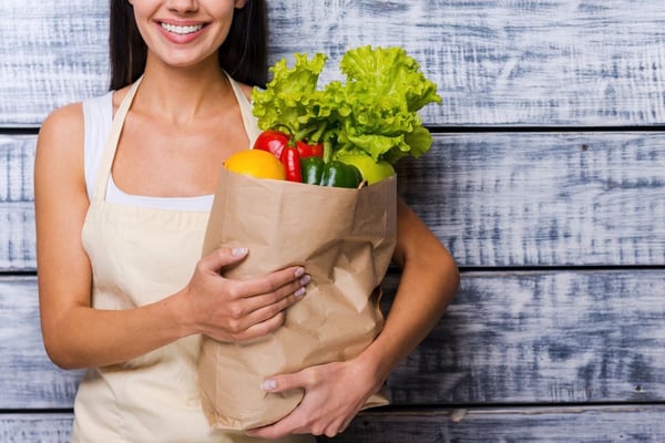 Woman in apron holding paper bag of fresh vegetables including lettuce, yellow pepper, and red peppers against wooden background