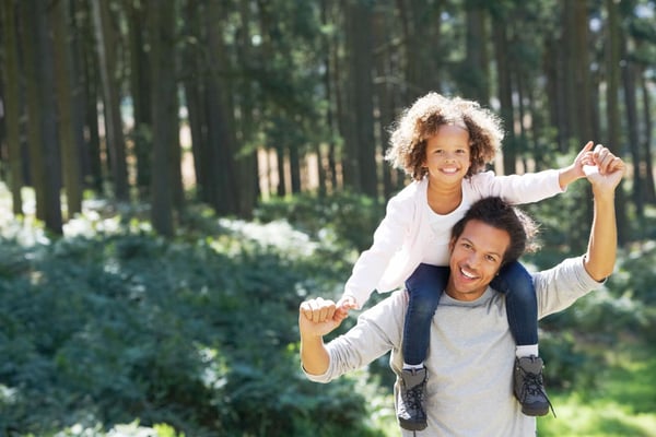 Father carrying young child on shoulders on a forest path lined with tall evergreen trees