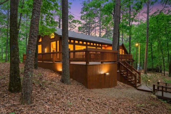 Cabin exterior lit up at twilight with warm glowing windows and wraparound deck surrounded by trees