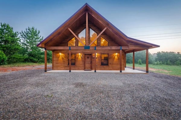 Modern wooden cabin with peaked roof, large windows, covered porch, and gravel driveway surrounded by forest