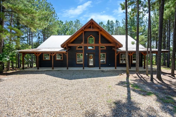 Modern log cabin with metal roof and covered porches surrounded by tall pine trees and gravel driveway