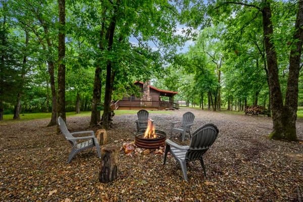Fire pit with Adirondack chairs surrounded by tall trees, brown cabin with red roof visible in background