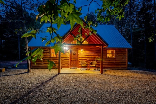 Log cabin exterior lit at night with porch light and chair, surrounded by trees